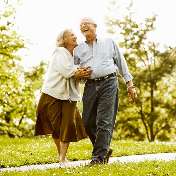 An elderly couple walks together outside on a sunny day, laughing and smiling, surrounded by green grass and trees.