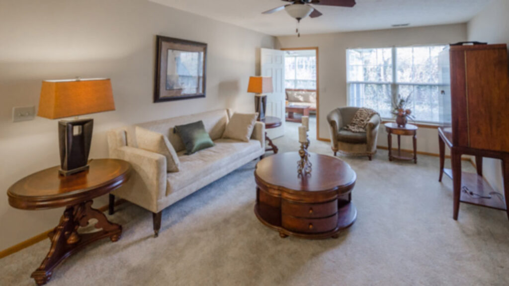 A cozy living room with beige carpet, a cream sofa, wooden tables, armchairs, lamps, and a large window letting in natural light. A door is open to a sunroom in the background.