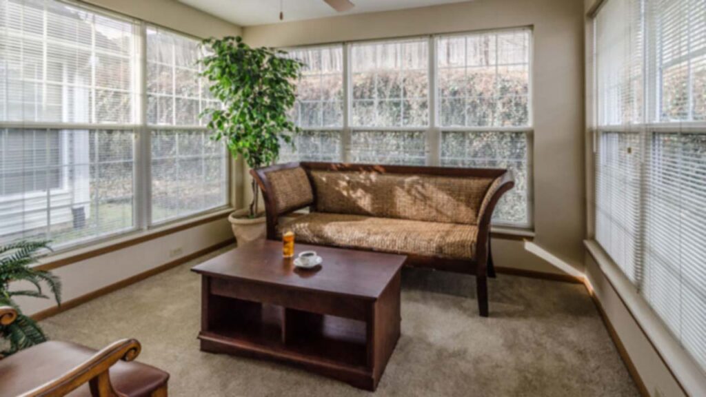 A sunlit room with large windows, a woven rattan-style loveseat, a wooden coffee table holding a cup and bottle, beige carpet, and a potted plant in the corner.