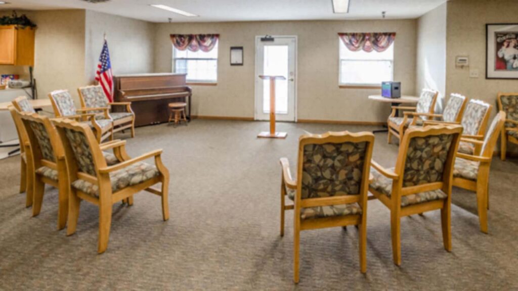 A room with a circle of wooden chairs, a piano, an American flag, a lectern, windows with patterned curtains, and beige carpeted floors, resembling a community meeting or group therapy space.