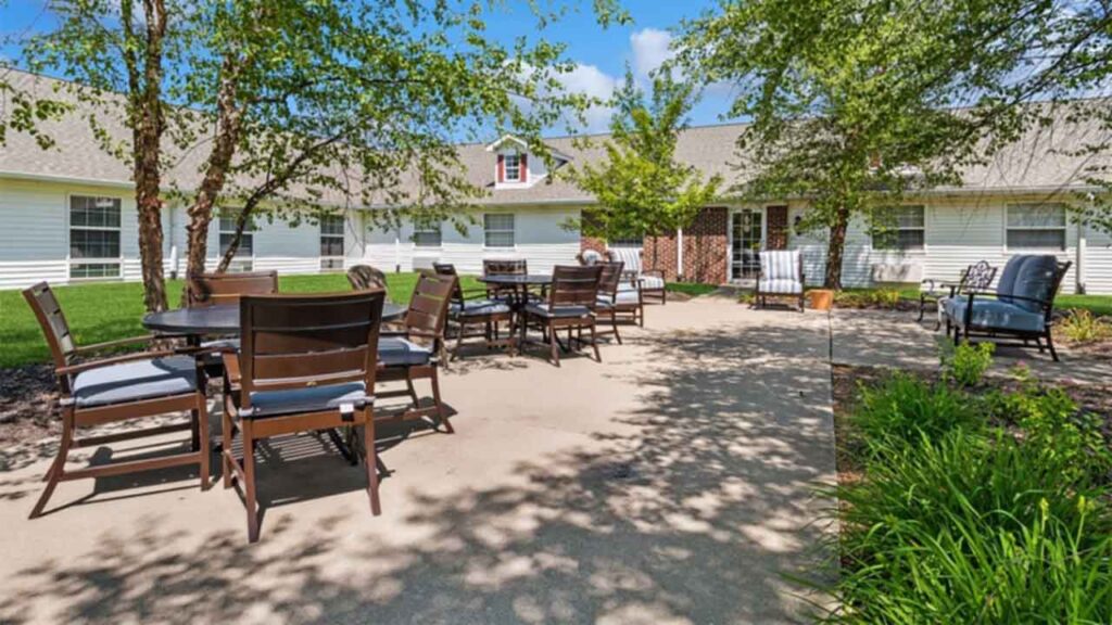 Outdoor patio with several round tables and cushioned chairs on a concrete walkway, surrounded by green grass, shrubs, and leafy trees in front of a white building with a gabled roof.