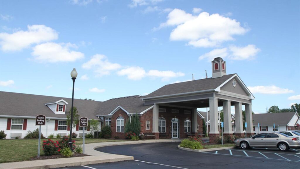 A single-story brick and white building with columns at the entrance, a small cupola on the roof, surrounding landscaping, parking spaces, and a partly cloudy sky above.