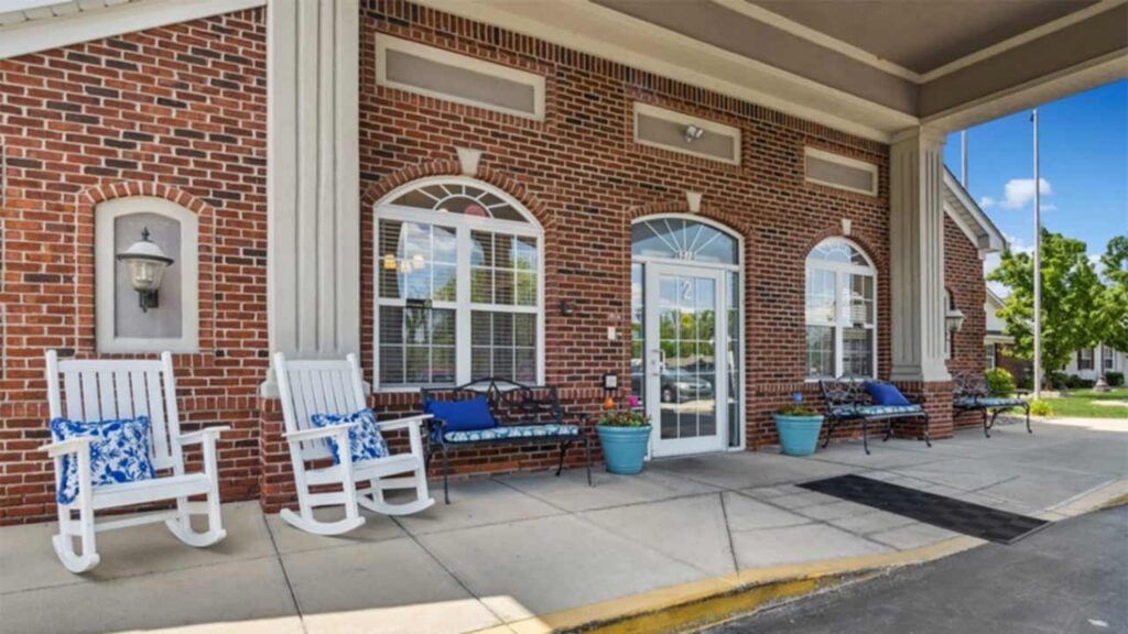 A brick building entrance with arched windows, white-framed glass doors, white rocking chairs with blue cushions, benches with pillows, and potted plants on a covered walkway.