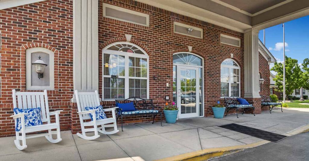 A brick building entrance with large windows, benches, rocking chairs with blue and white cushions, and potted plants on the sidewalk under a covered walkway.
