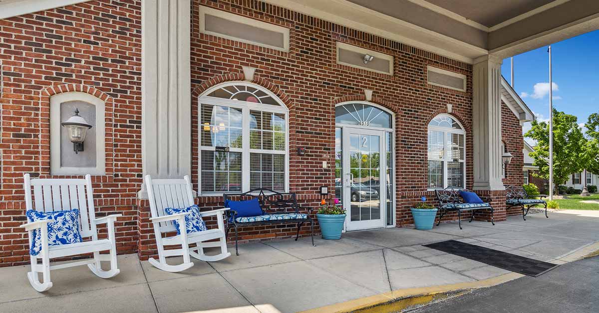 A brick building entrance with large windows, benches, rocking chairs with blue and white cushions, and potted plants on the sidewalk under a covered walkway.
