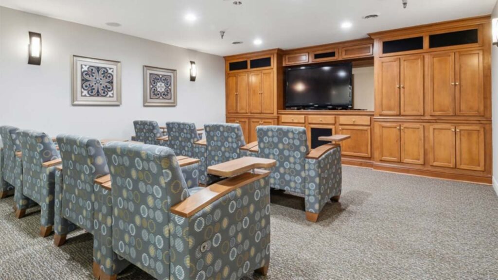 A small theater room with patterned armchairs in rows facing a large flat-screen TV set in wooden cabinetry; wall lights and framed art decorate the white walls.