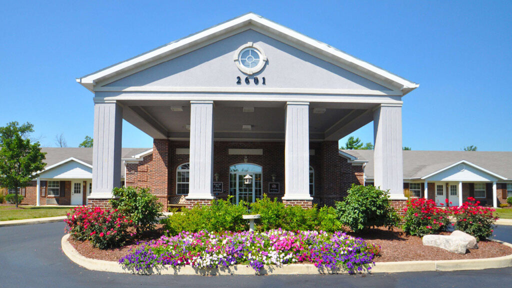 A single-story brick building with white columns and a covered entrance, surrounded by colorful flowerbeds and neatly trimmed bushes on a clear, sunny day.