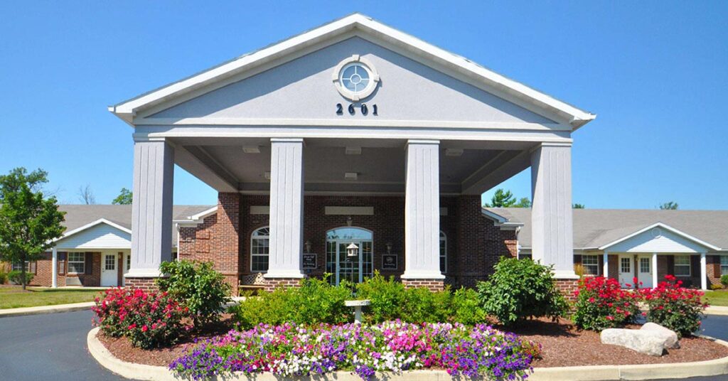A large brick building with white columns and a covered entrance surrounded by colorful flowers and neatly trimmed bushes, set against a clear blue sky. The number 2601 is displayed above the entrance.
