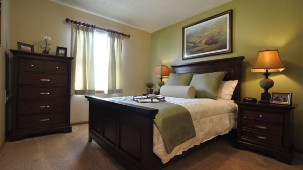 A cozy bedroom with a wooden bed, green and white bedding, matching nightstands with lamps, a dresser, a window with light curtains, and a framed painting on an olive green wall.