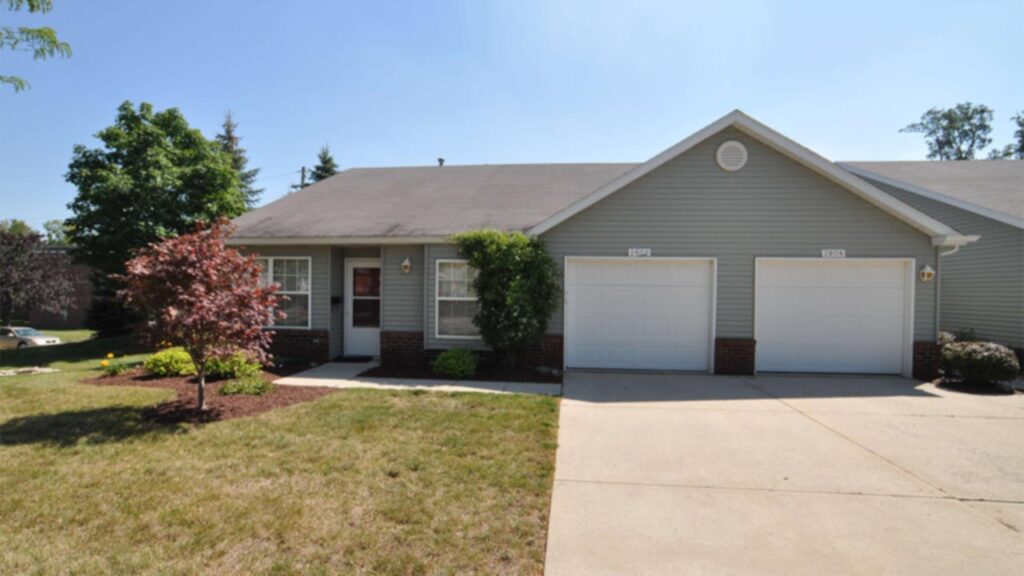 A single-story duplex with light gray siding, white trim, and two attached garages. There is a small front garden with shrubs and a red-leafed tree, and a concrete driveway leads up to the garages.