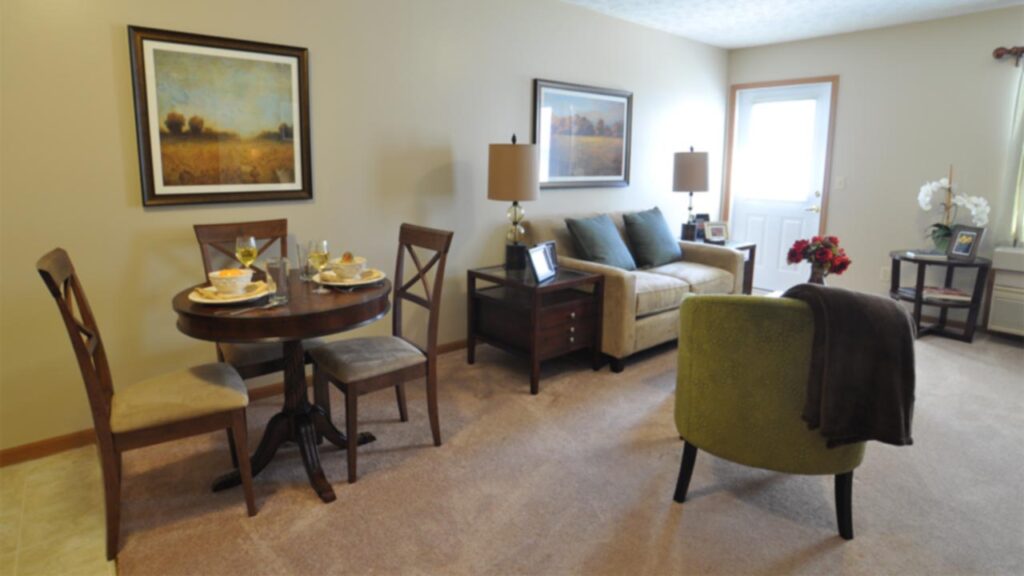 A cozy living and dining area with a round dining table set for three, a beige sofa, green armchair, side tables with lamps, framed artwork on the walls, and a door letting in natural light.