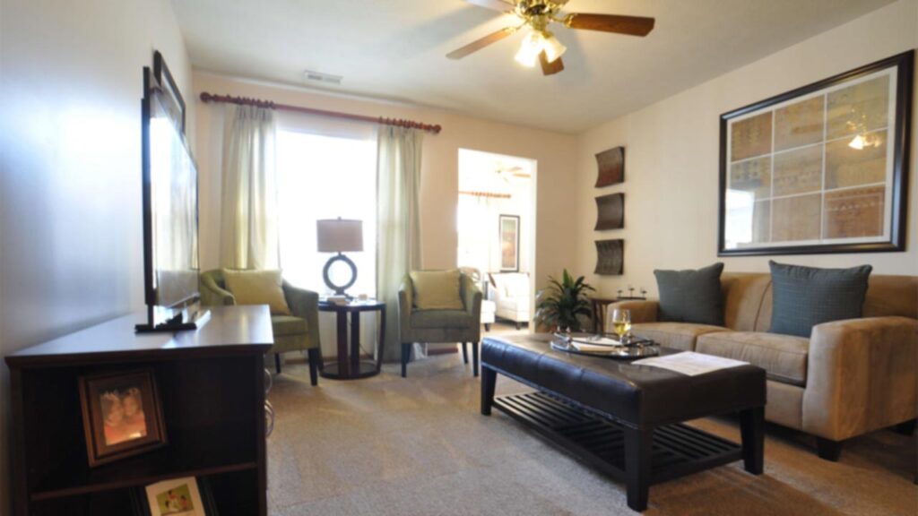 A bright living room with beige carpet, a tan sofa, two green chairs, a round table with a lamp, a cushioned coffee table, wall art, a TV on a stand, and a ceiling fan with lights. Sunlight shines through large windows.