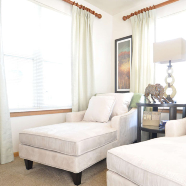 A bright living room with two white chaise lounge chairs, a side table with a lamp, books, and decorative animal figurines, and large windows with light curtains letting in natural light.