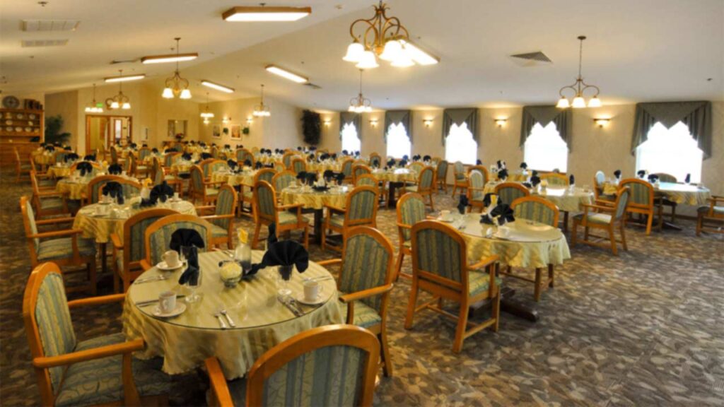 A spacious, well-lit dining room with many round tables covered in pale tablecloths, each set with cups, plates, and silverware. Wooden chairs with upholstered seats surround the tables. Chandeliers hang from the ceiling.