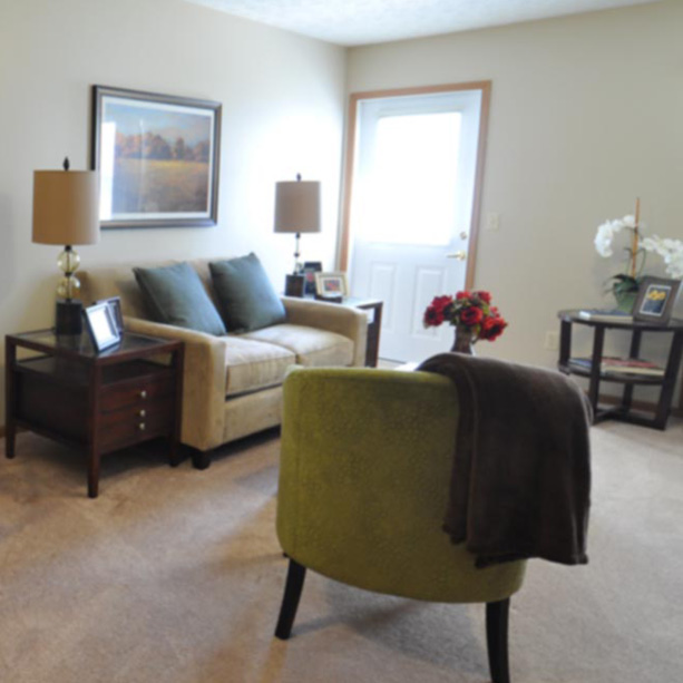 A cozy living room with a beige sofa, green armchair, side tables with lamps, framed artwork on the wall, a door letting in light, and decorative plants and flowers arranged around the room.