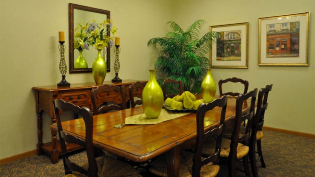 A traditional dining room with a wooden table and eight chairs, a sideboard with green vases and candlesticks, framed art on cream walls, and a large potted plant in the corner.
