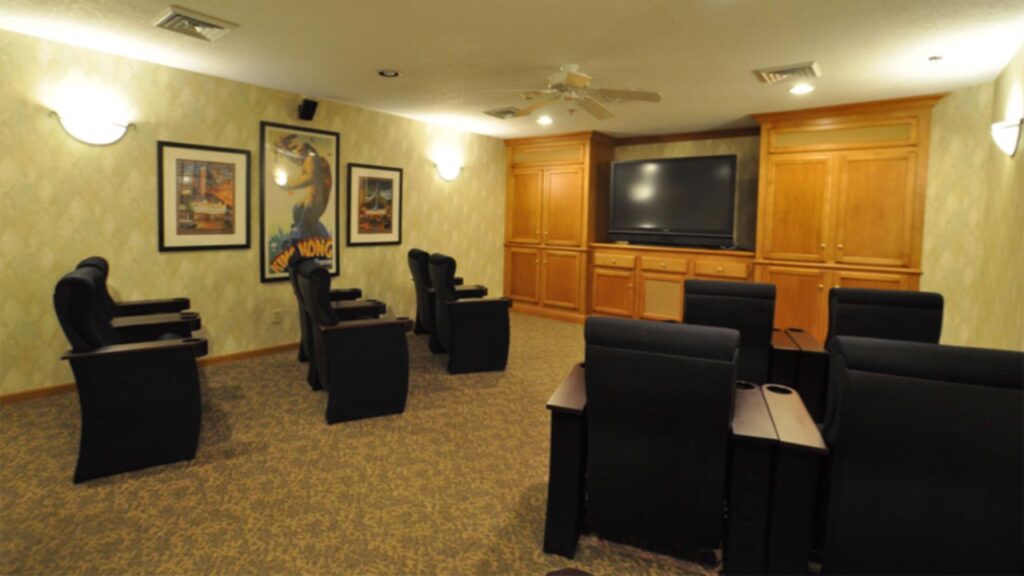 A small home theater room with eight black recliner chairs facing a large flat-screen TV, wood cabinets, framed movie posters on the walls, and carpeted flooring under warm lighting.