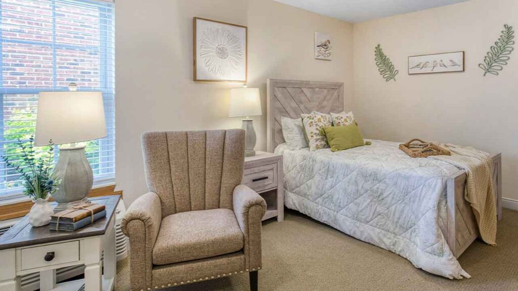A bedroom featuring an upholstered armchair, a side table with a lamp, and a bed with a patterned quilt and decorative pillows.