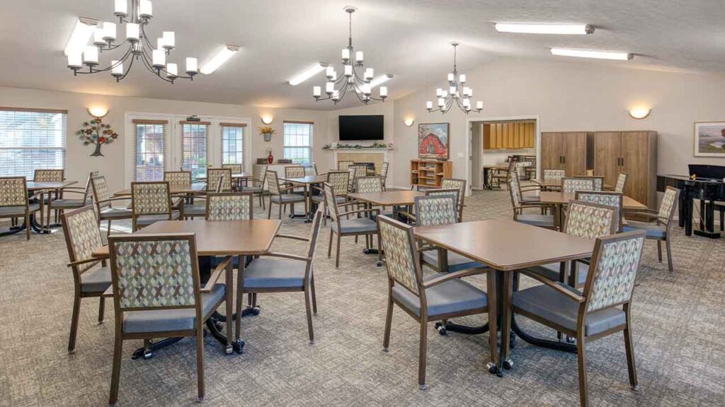 Dining room with many square tables and chairs arranged on a carpeted floor under chandeliers.