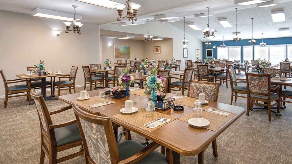 Dining room with multiple wooden tables set for meals, each featuring floral centerpieces, placemats, and place settings.