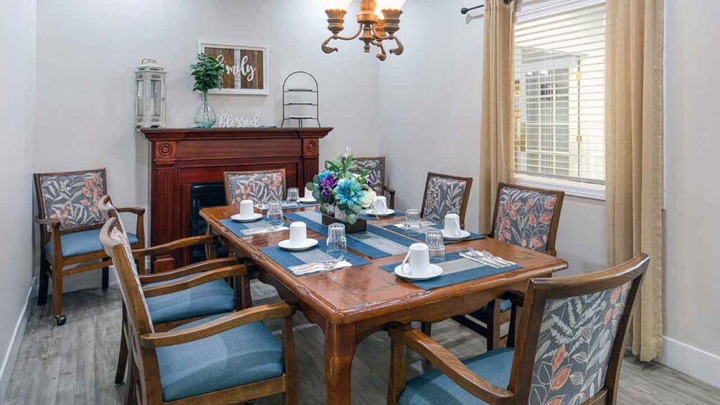 A wooden dining table set for six, surrounded by chairs with floral-patterned cushions and blue upholstery, in front of a fireplace with a mantel.