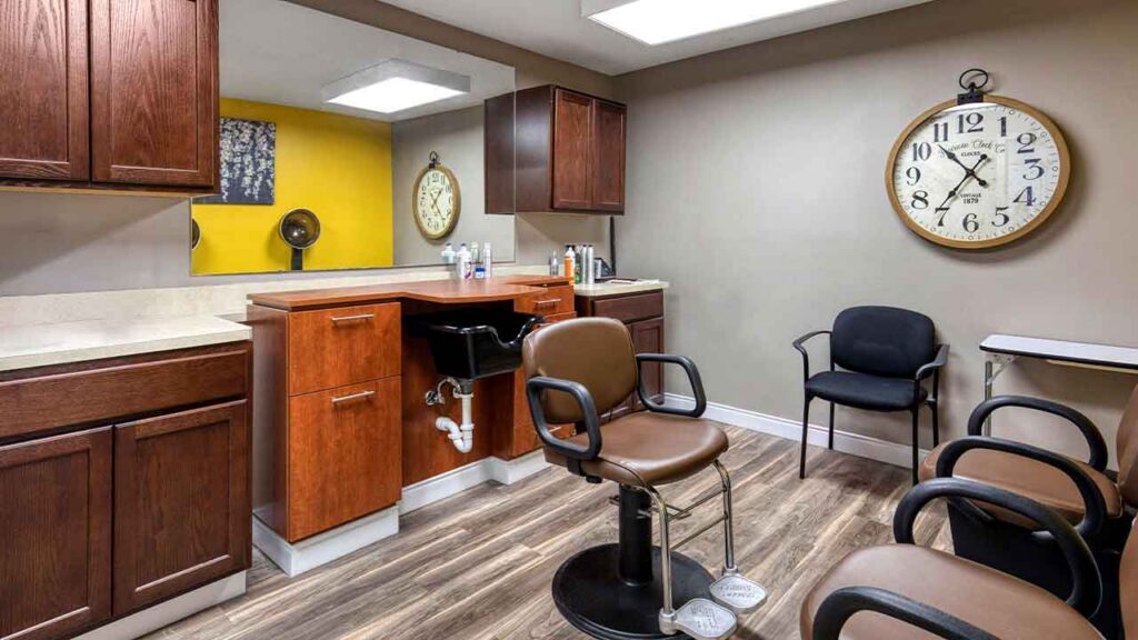 A hair salon station featuring a brown styling chair, a shampoo sink, and two additional chairs for waiting, with a large wall clock displayed prominently.