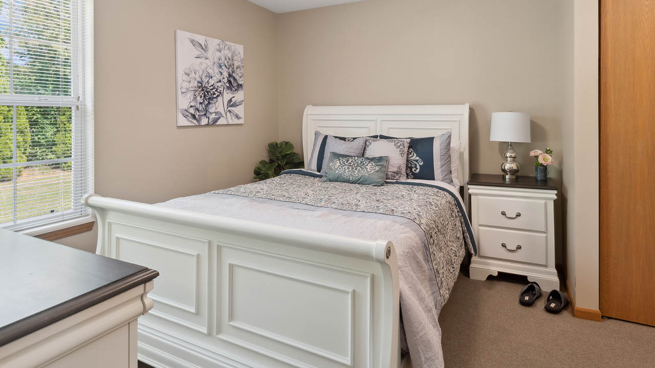 A cozy bedroom with a white bed frame, floral bedding, and matching nightstand. A lamp and decor items sit on the nightstand. A large window lets in natural light, and a floral painting hangs above the bed.