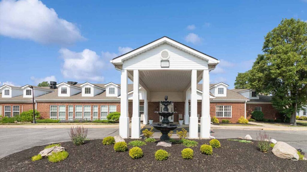 A brick building with white trim and dormer windows, featuring a covered entrance supported by columns. A tiered black fountain and landscaped shrubs are in the foreground under a blue sky with scattered clouds.