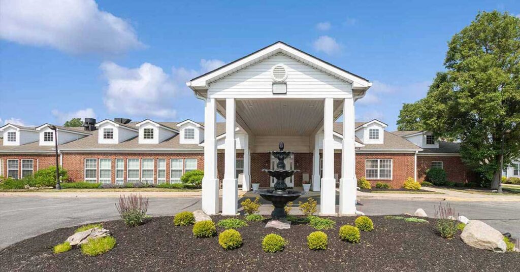 A red-brick building with multiple windows and a white portico entrance. A black, tiered fountain sits in front, surrounded by neatly trimmed bushes and landscaping under a blue sky with scattered clouds.