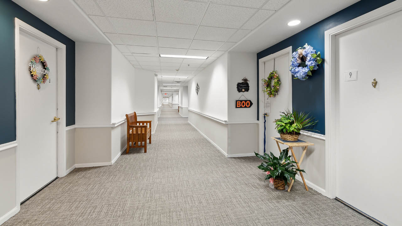 A well-lit hallway in an apartment or assisted living facility, with decorated doors, potted plants, and a bench along the wall. Door number 9 is visible with a wreath and a "BOO" sign.