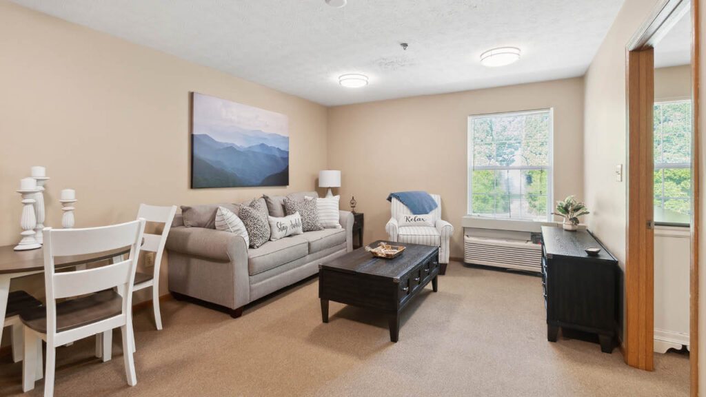 A cozy living room with beige walls, tan carpet, a gray sofa, white armchair, black coffee table, and a TV stand. A dining table with white chairs is in the corner. Large windows let in natural light.