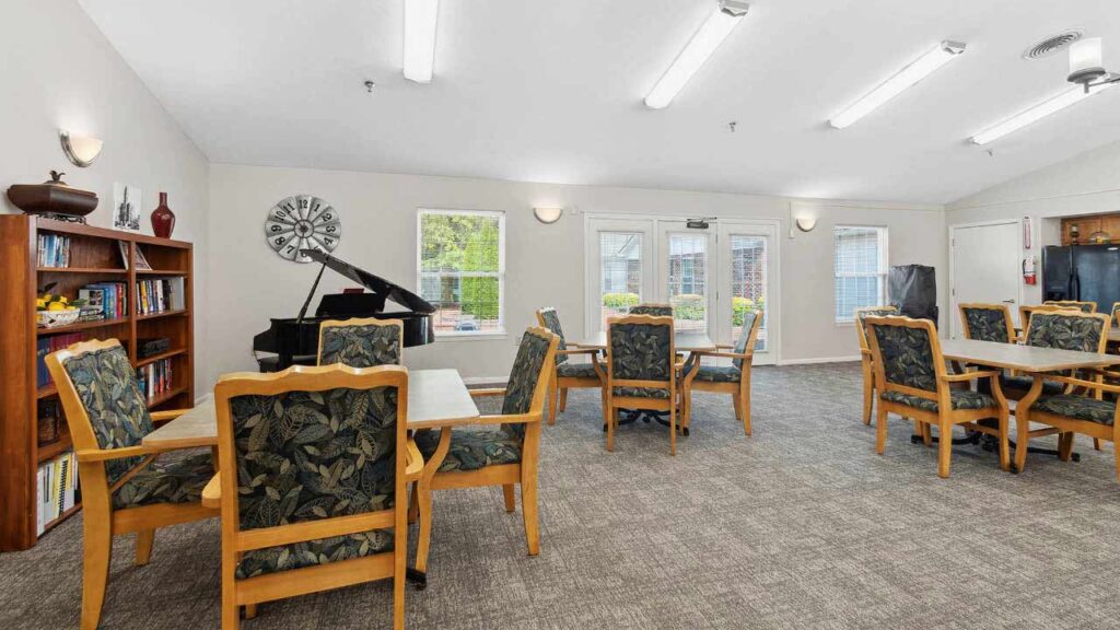 A spacious, well-lit common room with tables and chairs, a black grand piano, bookshelves, and large windows letting in natural light. The décor is cozy, with plants and wall art adding a welcoming touch.