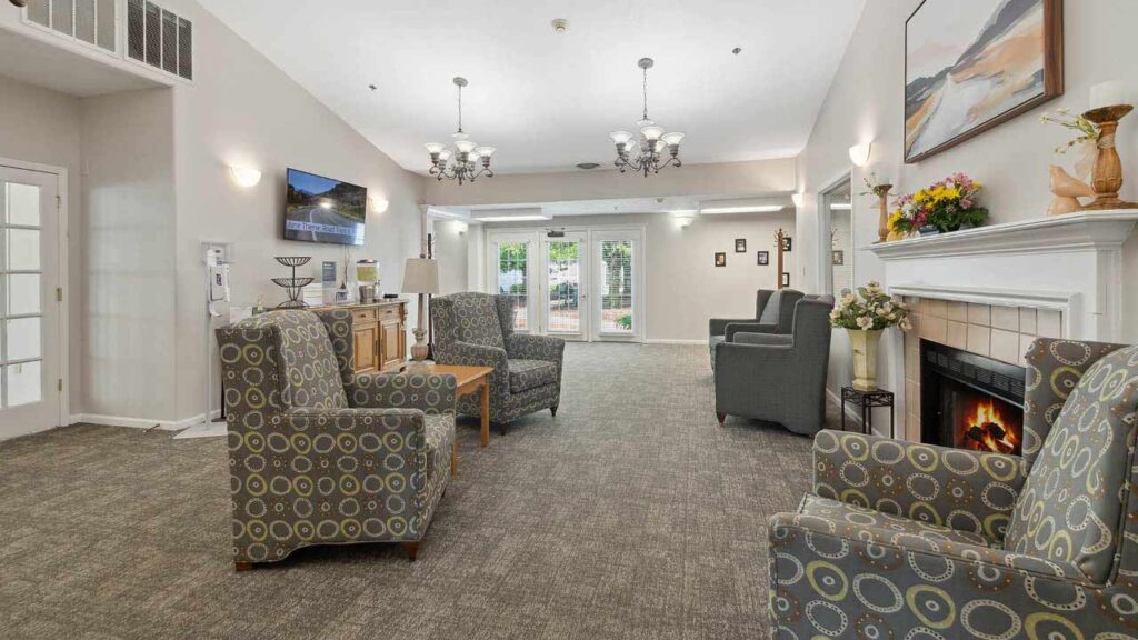 A cozy, well-lit living room with patterned armchairs, a lit fireplace, wall art, a TV, and large windows letting in natural light. Chandeliers hang from the ceiling, and flowers decorate the mantle.