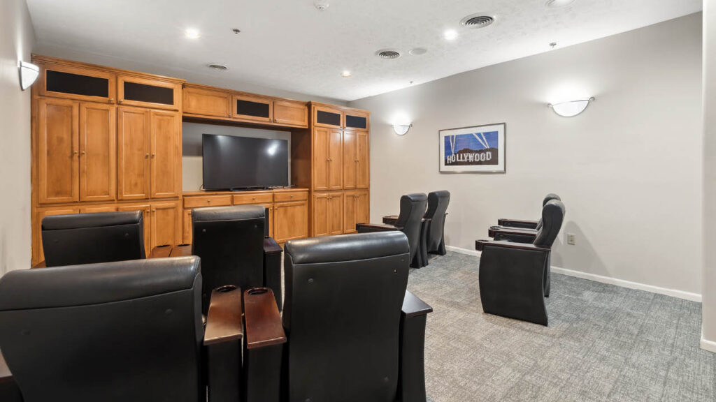 Small theater room with black leather chairs facing a flat-screen TV on a wooden entertainment center; gray carpet, beige walls, wall lights, and a framed 