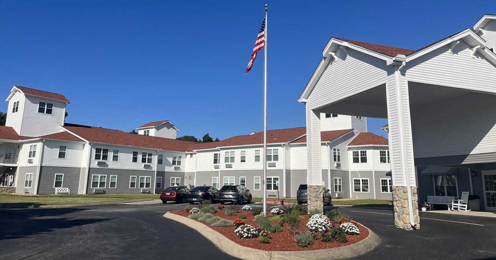 A white, two-story building with a red roof, a covered entrance, and a circular driveway with landscaped flowers and an American flagpole in front; several cars are parked along the drive under a clear blue sky.