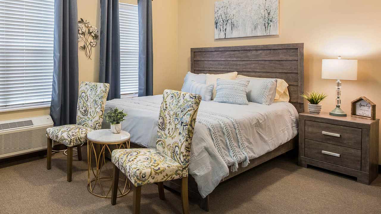 A cozy bedroom with a wooden bed, light bedding, two patterned chairs around a small round table, a nightstand with a lamp and plant, and windows with blue curtains. Artwork hangs above the bed.