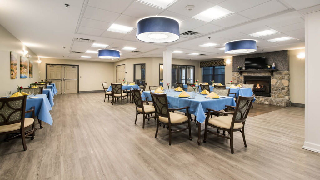 Spacious, well-lit dining room with wooden floors, square tables covered in blue tablecloths, neatly set with napkins and silverware, cushioned chairs, and a stone fireplace in the background.