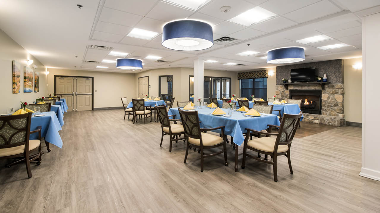 Spacious, well-lit dining room with wooden floors, square tables covered in blue tablecloths, neatly set with napkins and silverware, cushioned chairs, and a stone fireplace in the background.