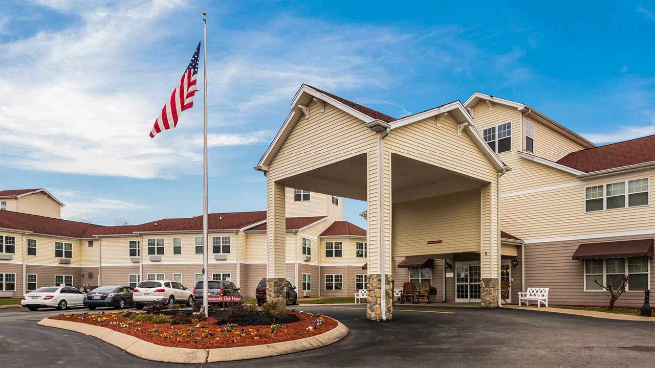 A beige, two-story building with a covered entrance and a U.S. flag at half-staff stands under a blue sky. Several cars are parked in front, and landscaped flower beds border the driveway.