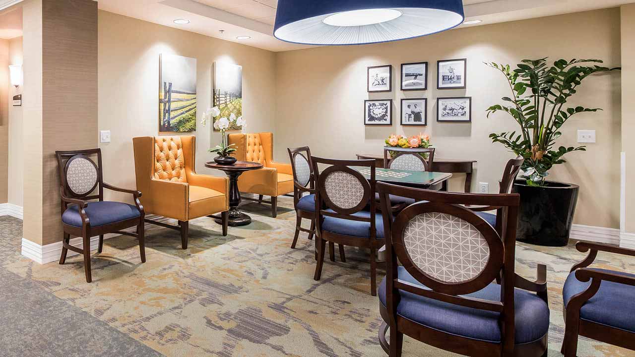 A cozy lounge area with orange armchairs, dark wood tables and chairs, wall art, and a large potted plant on patterned carpet under a modern ceiling light.