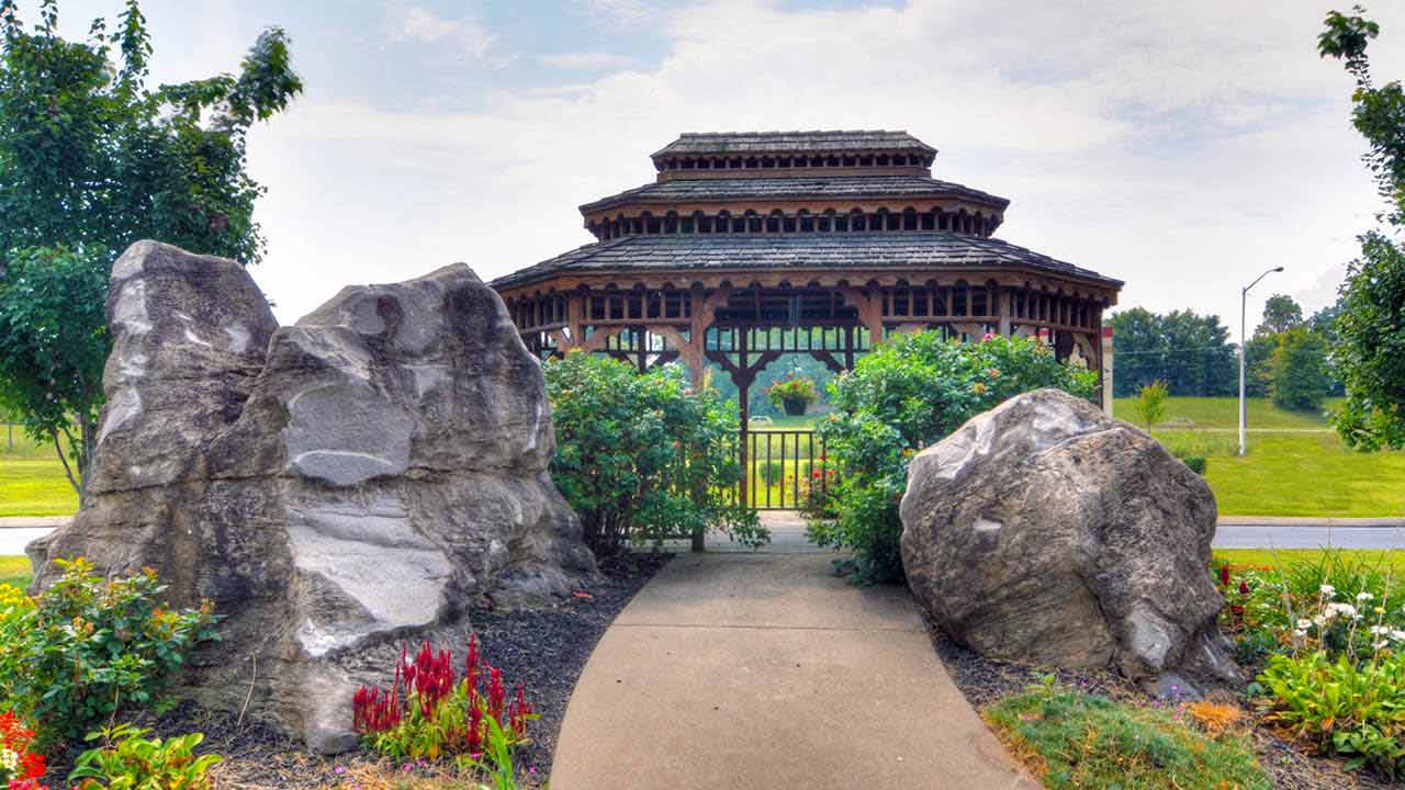 A paved path lined with flowers and bushes leads between two large rocks to a wooden gazebo with a tiered roof, set in a grassy park with trees in the background.