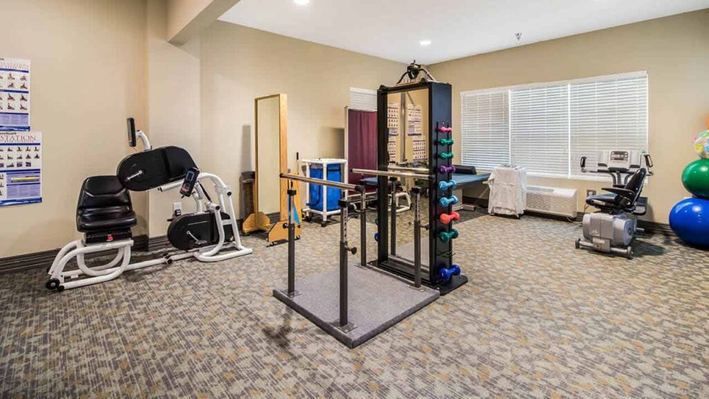 A gym room with exercise equipment including a stationary bike, a weight machine, free weights on a rack, therapy balls, and charts on the wall. The room has carpeted floors and large windows with blinds.