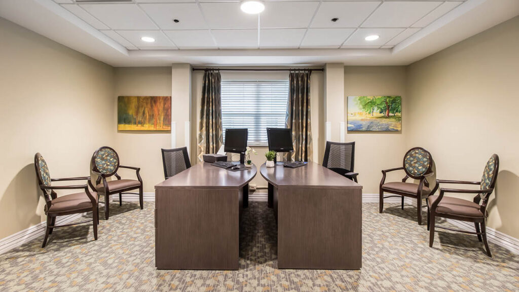 A modern office with two wooden desks placed in an L-shape, each with a computer monitor and chairs. Four guest chairs are arranged along the walls, and two landscape paintings hang on the beige walls. Curtains frame a window.
