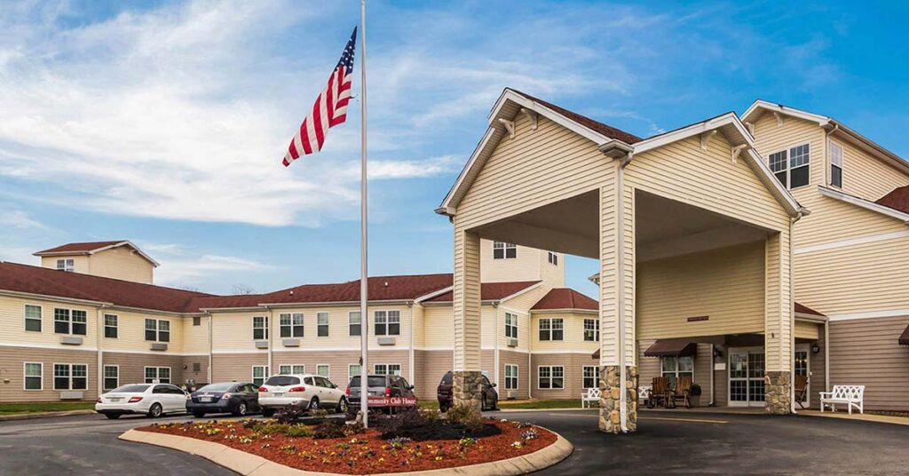 A beige two-story building with an American flag in front, a covered entrance, landscaped flowerbeds, and several parked cars, under a partly cloudy blue sky.