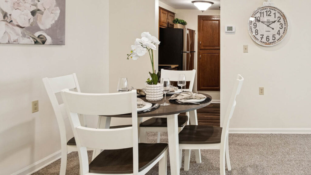 A dining area with a round table set for four, white chairs, a floral centerpiece, and neutral decor. A kitchen with wooden cabinets and a wall clock are visible in the background.