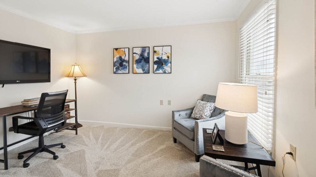A bright home office with a desk, chair, TV on the wall, lamp, and three abstract paintings. A cozy armchair and side table with a lamp sit by a large window with white blinds, letting in natural light.