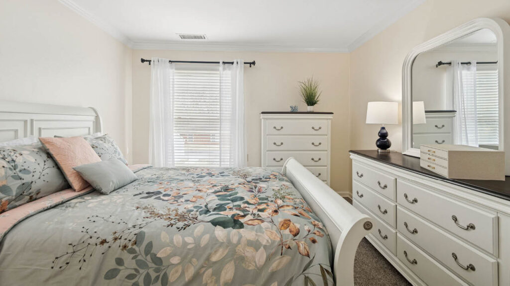 A bright bedroom with a white sleigh bed, floral bedding, two dressers, a mirror, a table lamp, decorative boxes, a potted plant, and large windows with white curtains letting in natural light.