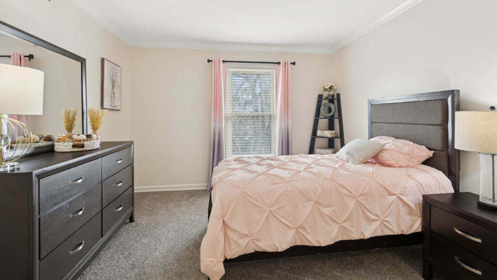 A cozy bedroom with a bed covered in a pink comforter, two bedside lamps, a dresser with a mirror, a window with pink curtains, and a black corner shelf with decorative items.