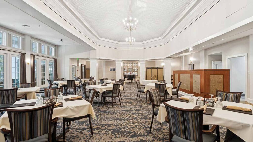 A spacious, well-lit dining room with round tables covered in cream tablecloths, set with glasses and napkins. Elegant chairs surround each table, and large windows let in natural light. Chandeliers hang from a high, vaulted ceiling.