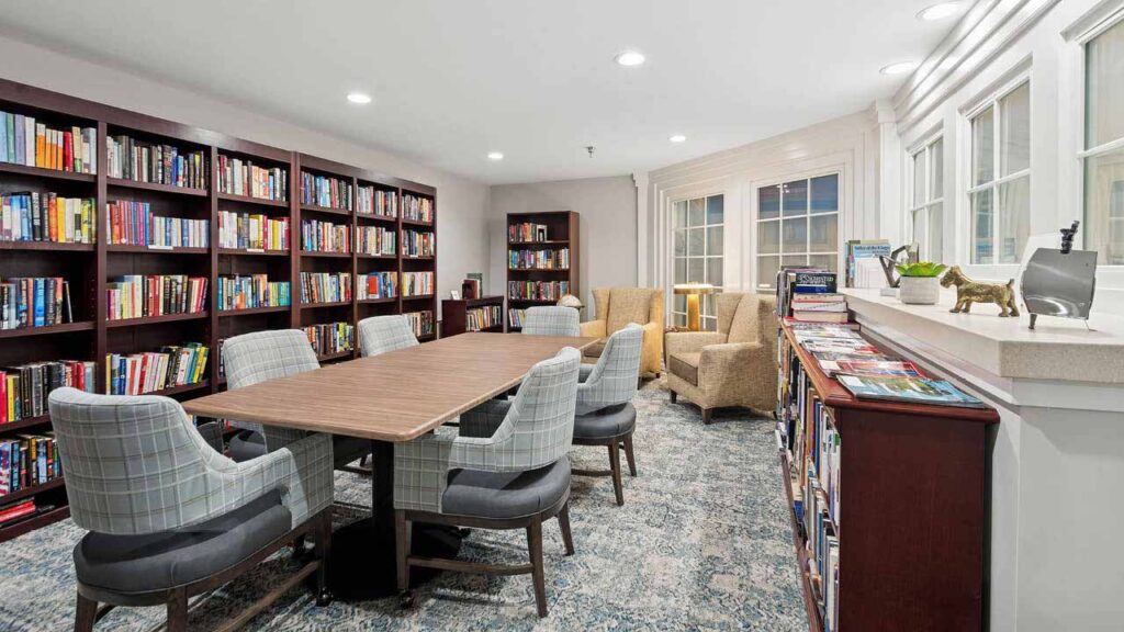 A cozy library room with bookshelves lining the walls, a long wooden table surrounded by cushioned chairs, two armchairs by large windows, and a side shelf holding magazines and decorative items.